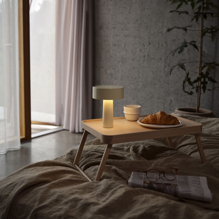 Eine tragbare Lampe im modernen Stil steht auf einem hellen Holztablett mit einem Croissant und einer Tasse auf einem Bett in einem minimalistisch eingerichteten Schlafzimmer mit grauer Wand, großen Fenstern und gedämpftem Licht.