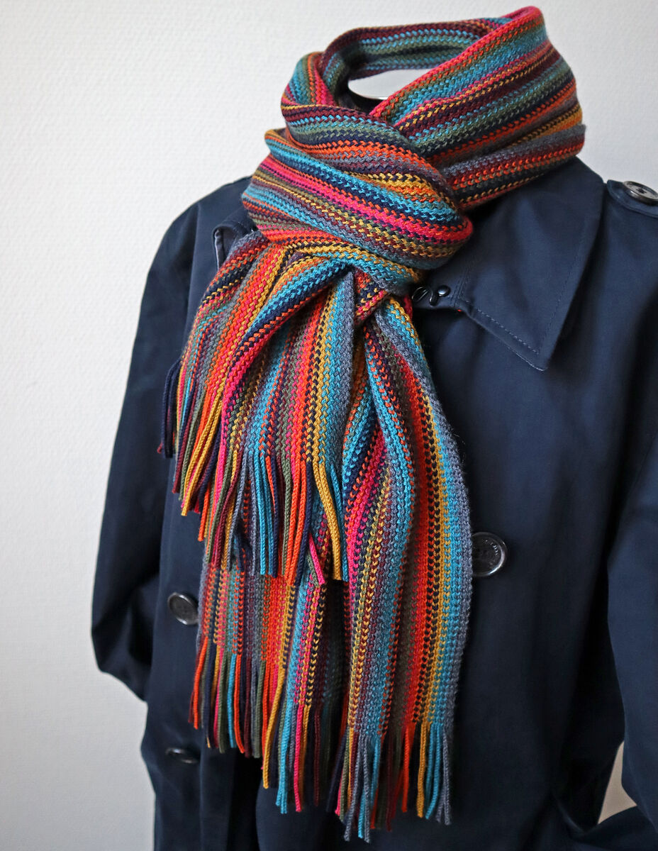 Bunter Merinoschal mit Streifen in Blau, Orange, Rot, Grün und Gelb, kunstvoll um den Hals einer dunklen Mantelpuppe gewickelt. Der Schal wirkt kuschelig und stilvoll und eignet sich als modisches Accessoire für Herbst und Winter.