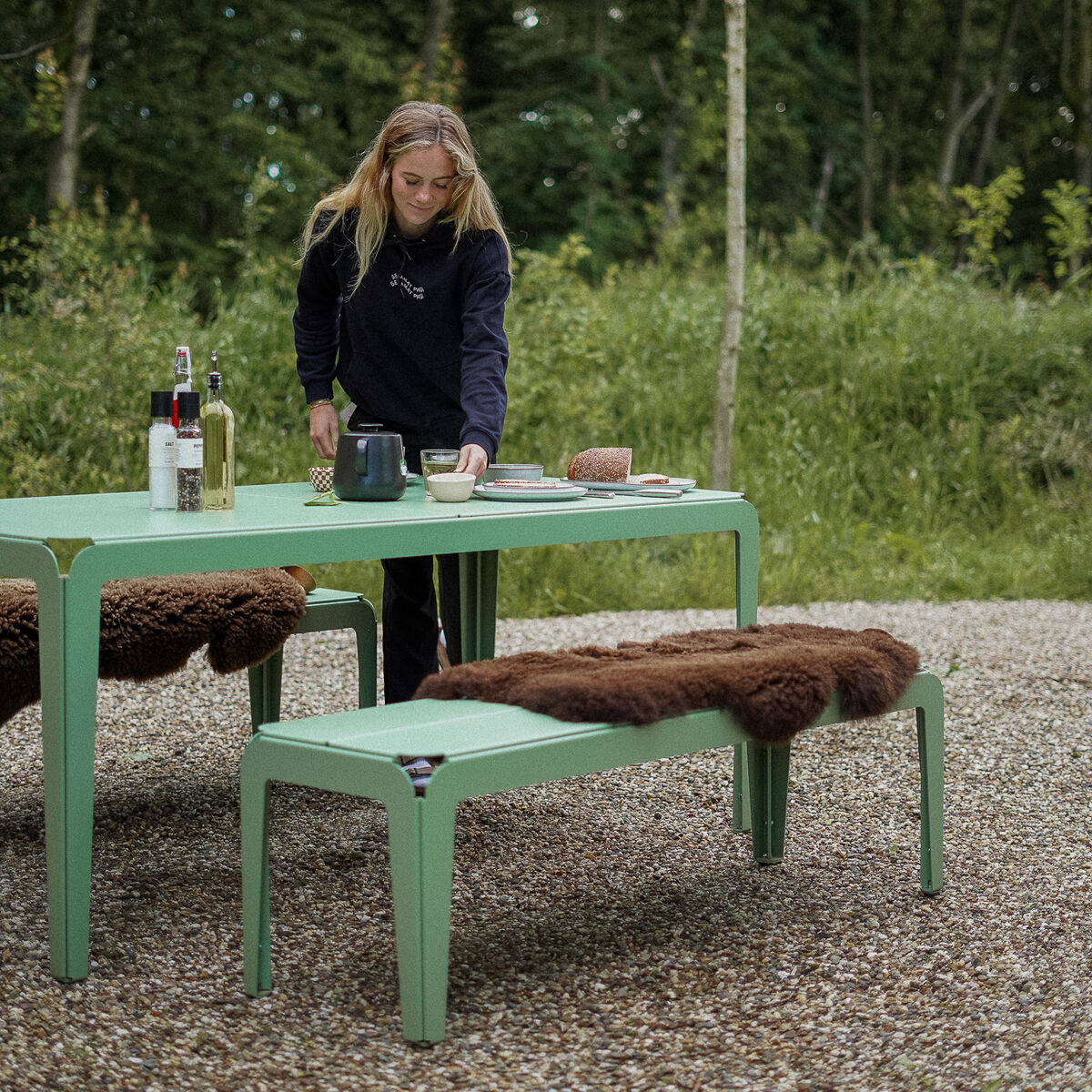 Eine Frau deckt einen grünen Gartentisch für ein Picknick im Freien, während auf der grünen Gartenbank ein braunes Schafsfell mit Lederschlaufe als Sitzauflage liegt. Im Hintergrund befindet sich eine natürliche, bewaldete Umgebung.