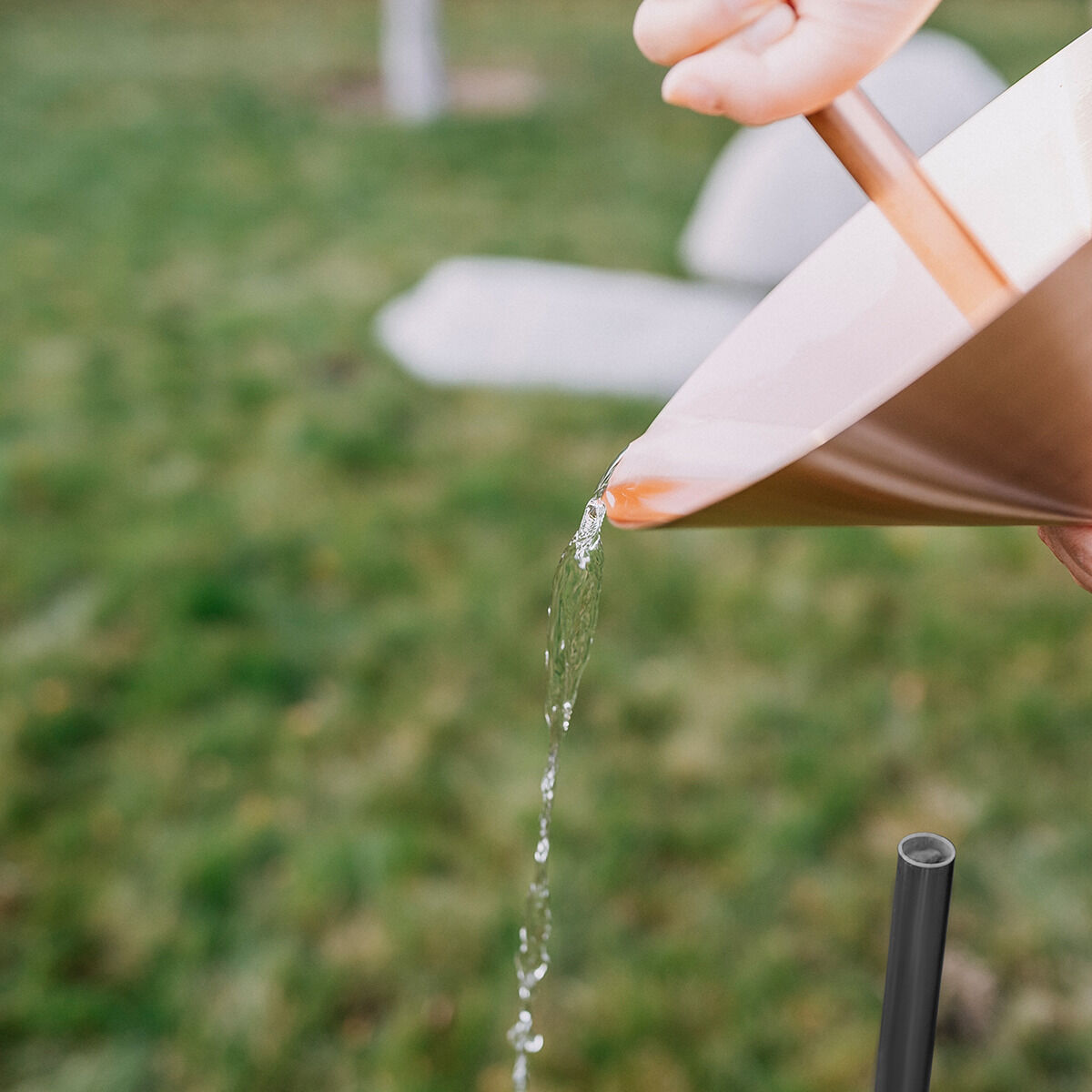 Nahaufnahme eines Regenmessers aus Kupfer der Marke Fischer Manufaktur, der von einer Hand geleert wird. Das glänzende Kupfergefäß gießt Wasser aus, im Hintergrund ist eine unscharfe grüne Wiese und Gartenfläche zu sehen.