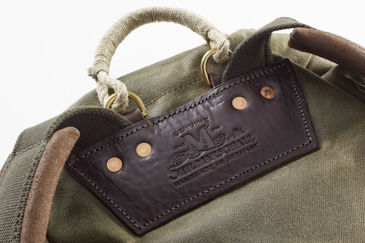 Oberer Teil eines robusten olivgrünen Rucksacks mit braunen Lederdetails und beigem Seilstangengriff. Rindslederaufkleber mit der Prägung 'Handcrafted in Germany'.