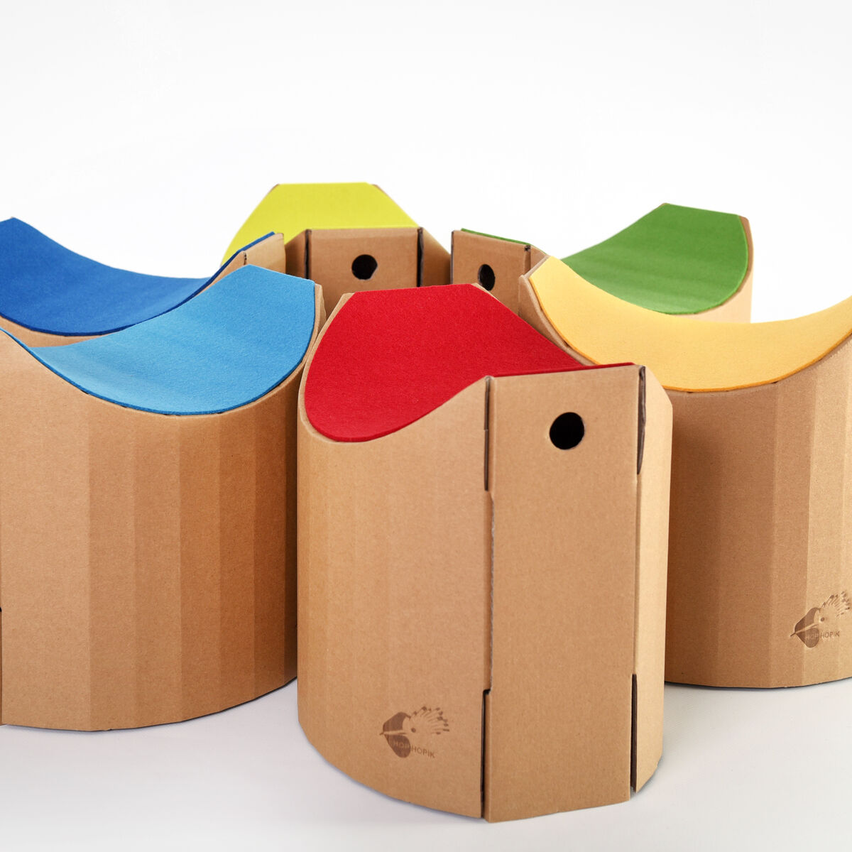 Eine Gruppe von Papphockern namens »Lollo« in verschiedenen Farben (blau, rot, grün, gelb) mit einem naturbraunen Kartonbasis. Der Hocker hat eine gebogene Sitzfläche und ist modular aufgebaut.