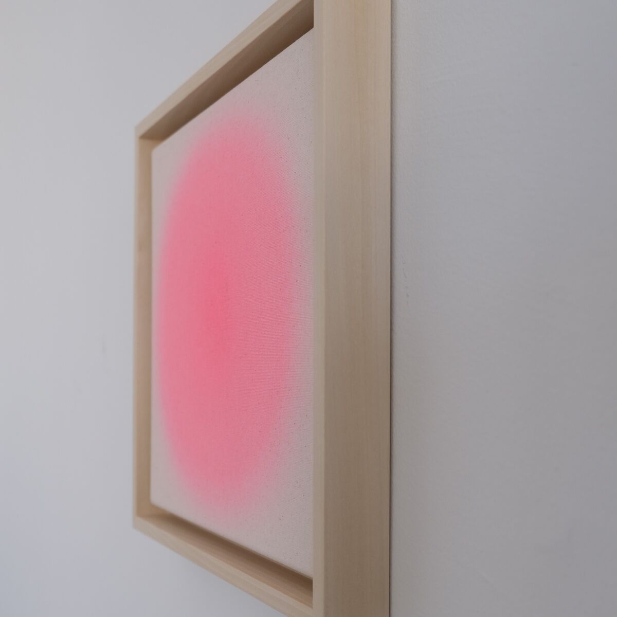 Gerahmtes quadratisches Kunstwerk mit sanftem pinkfarbenem Farbverlauf auf weißem Grund, aufgehängt an einer weißen Wand. Moderner minimalistischer Stil mit hellem Holzrahmen.