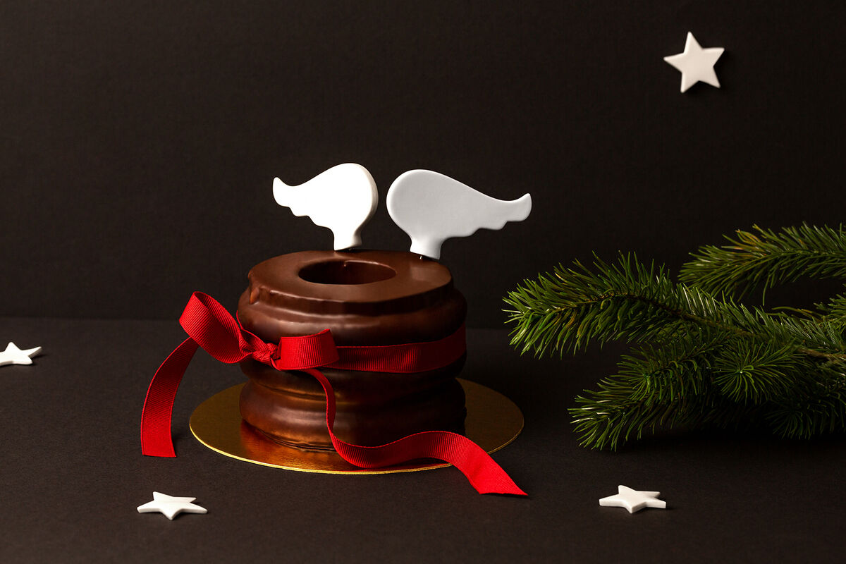 Eine Torte mit Schokoladenüberzug, dekoriert mit weißen Flügelsteckern aus Porzellan und einer roten Schleife. Umgeben von Tannenzweigen und weißen Sternen auf schwarzem Hintergrund.