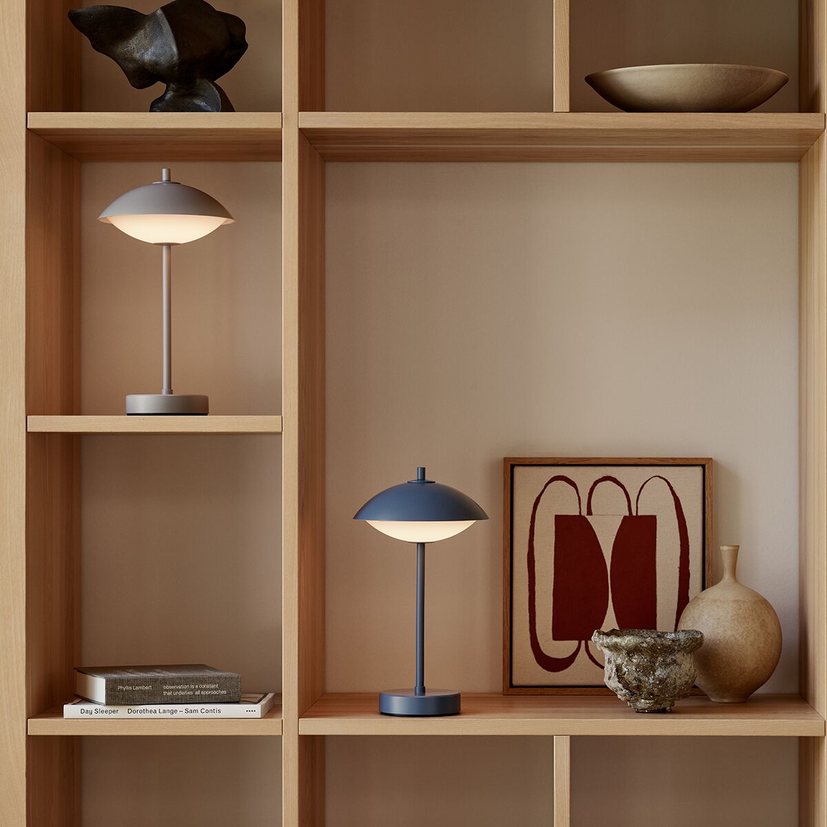 Auf einem hellen Regal stehen eine tragbare Lampe »Clam™« von Fritz Hansen in Grau, eine weitere identische Lampe, Kunstobjekte, Bücher, eine Vase und ein modernes abstraktes Bild. Die Szene wirkt aufgeräumt, modern und wohnlich.