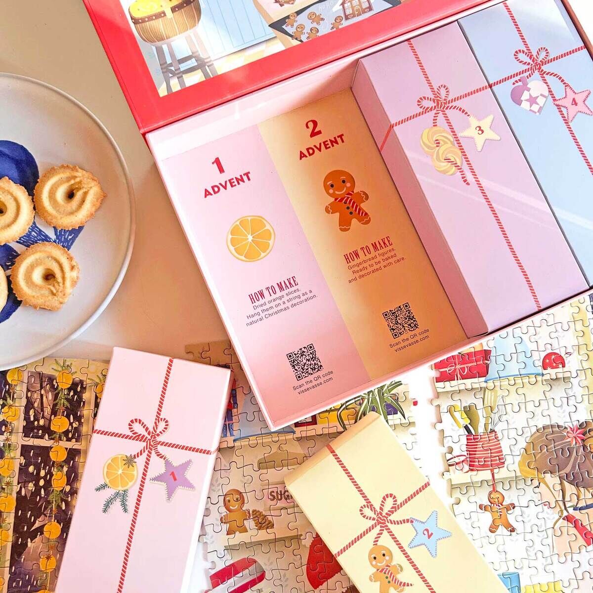 Der Puzzle-Adventskalender »Sweet Christmas« von VISSEVASSE zeigt mehrere pastellfarbene Geschenkboxen im geöffneten Karton und ein weihnachtliches Puzzle mit 1000 Teilen. Daneben liegt ein Teller mit Gebäck. Alles ist liebevoll und festlich gestaltet.