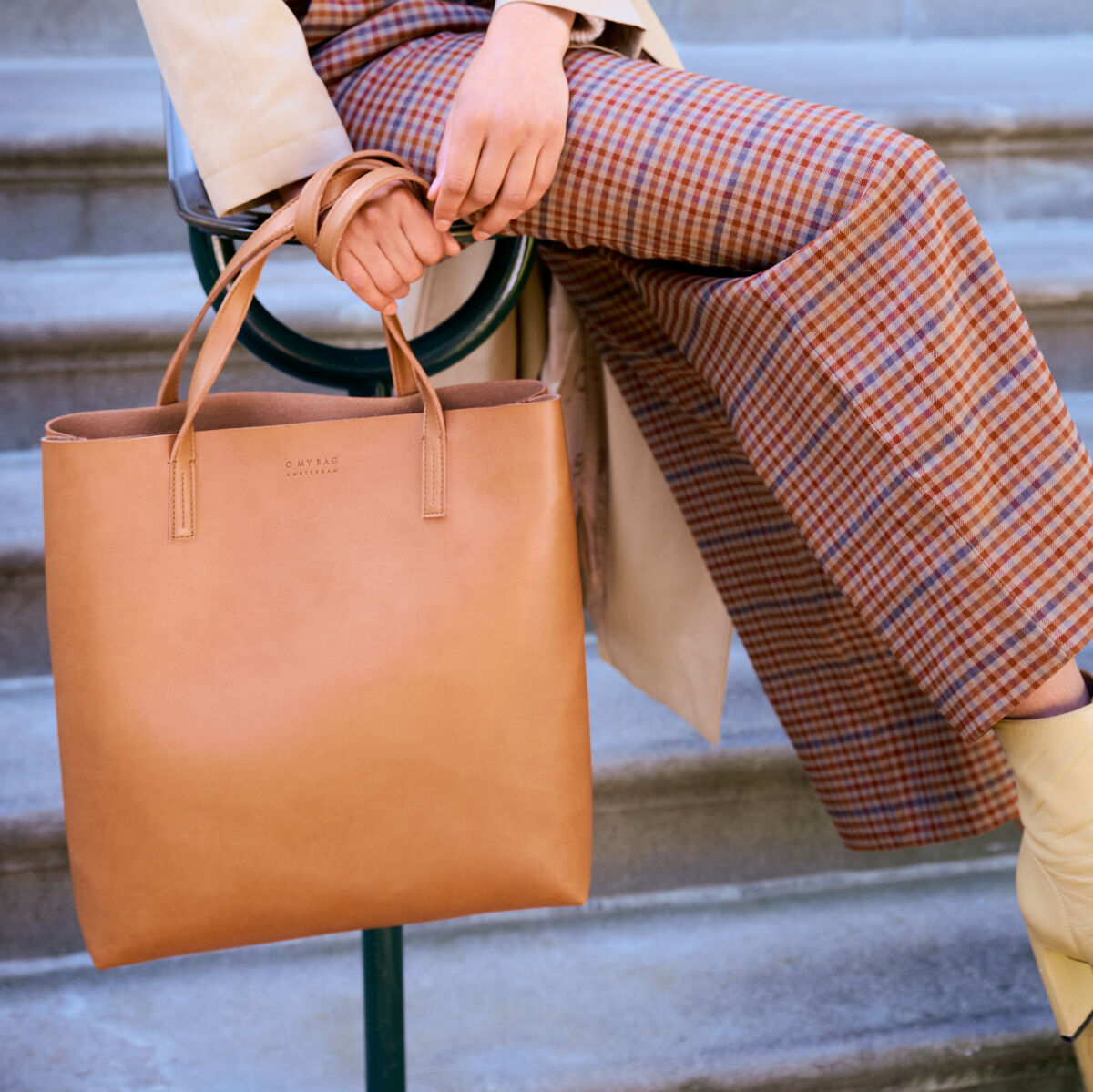 Eine Frau in stilvoller Kleidung sitzt auf einer Treppe und hält eine Shopper-Tasche »Georgia« aus braunem Leder. Sie trägt karierte Hosen und einen beigen Mantel.