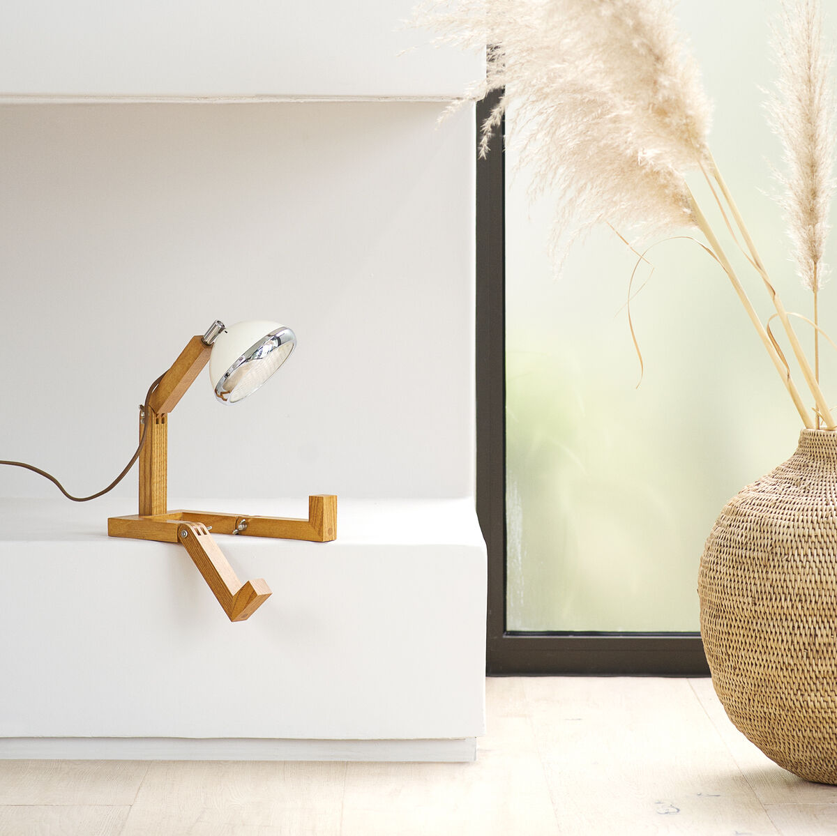 Bild einer modernen Tischleuchte aus Holz mit einem minimalistischen Design. Die Lampe steht vor einem großen Fenster und daneben ist eine dekorative Vase mit Pampasgras. Der Raum ist hell und minimalistisch eingerichtet.