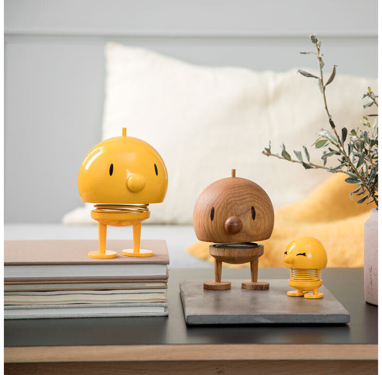 Holzfiguren der Marke Hoptimist Bumble aus Eichenholz. Drei Figuren in verschiedenen Größen und Farben, überwiegend gelb und naturfarben, auf einem Tisch in einem stilvoll eingerichteten Raum mit Büchern und Pflanzen im Hintergrund.