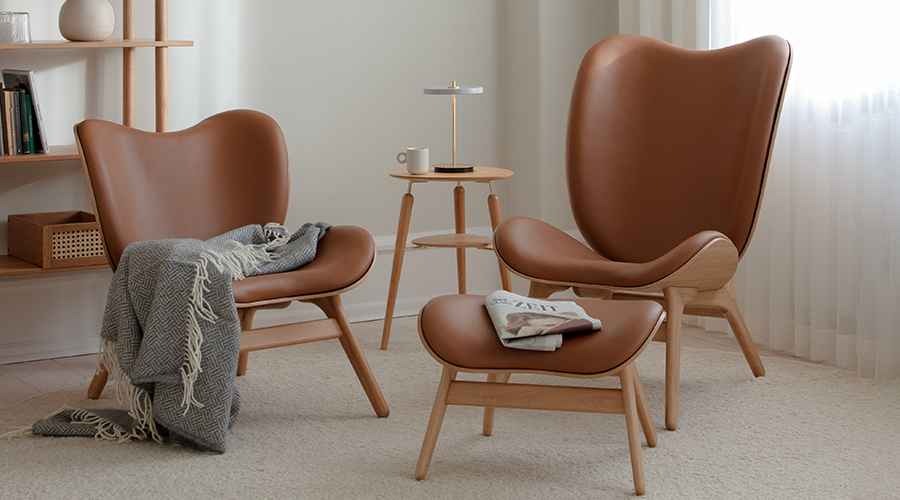 Zwei braune Ledersessel mit Holzrahmen, ein passender Hocker mit Zeitung, ein kleiner runder Beistelltisch mit Lampe und Tasse, graue Decke auf einem Sessel