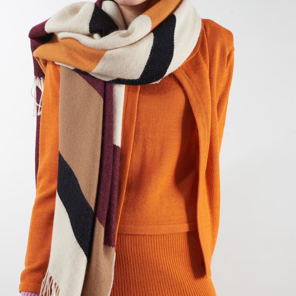 Eine Person trägt ein Cape aus Alpaka-Wolle in warmen Herbsttönen. Der Schal ist in Orange, Braun, Beige, Weiß, Schwarz und Bordeaux gehalten und sieht weich und bequem aus.