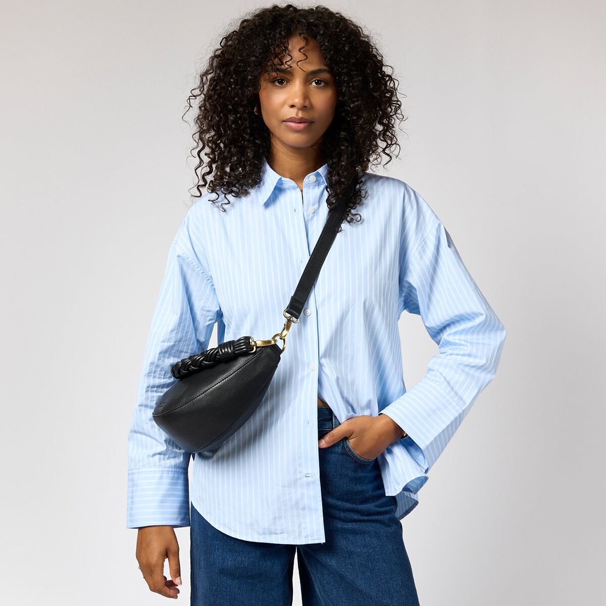 Eine Frau trägt eine schwarze Crossbody-Tasche »Leo« von O My Bag aus Kernleder quer über der Brust. Sie trägt ein hellblau-weiß gestreiftes Hemd und dunkelblaue Jeans. Der Hintergrund ist neutral hellgrau, die Tasche wirkt hochwertig mit goldfarbenem Detail am Riemen.