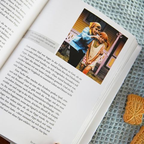 Ein aufgeschlagenes Buch zeigt einen Text auf der linken Seite und ein Foto von zwei Mädchen, die draußen vor einem gelben Haus stehen. Das Buch liegt auf einem strukturierten hellblauen Tuch und daneben liegt ein Lebkuchen-Plätzchen in der Form eines Herzens.