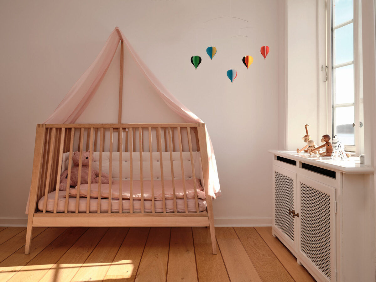 Ein Babyzimmer mit einem Holzgitterbett, das mit rosa Bettwäsche ausgestattet ist. Darüber hängt ein Mobile mit fünf bunten Heißluftballons. Links im Bett liegt ein Kuscheltier. Rechts steht ein weißer Schrank unter einem Fenster mit Sonnenlicht.