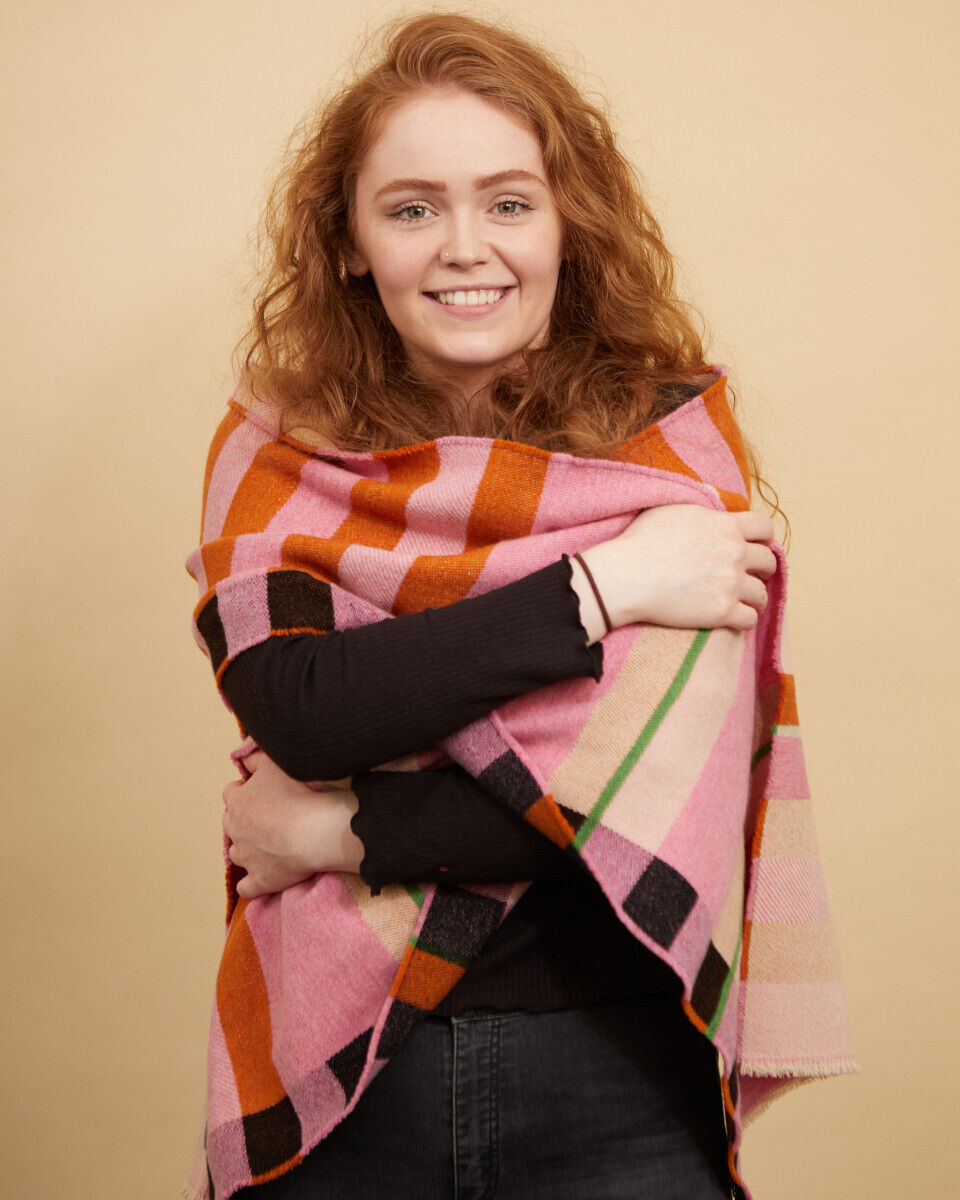 Eine Frau trägt einen farbenfrohen, gestreiften Schal bzw. Wrap im Stölzl-Bauhaus-Design. Die Farben des Schals umfassen Rosa, Orange, Schwarz, Grün und Rot. Das Design wirkt warm und stilvoll, ideal für kühle Jahreszeiten.