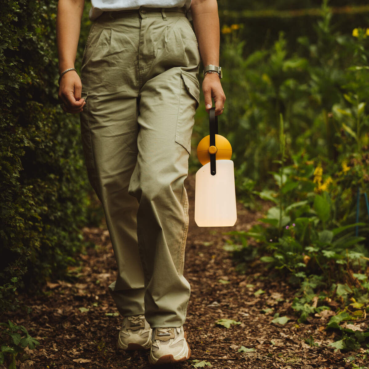 Person, die eine Guidelight Lampe in einem grünen Gartenweg trägt, umgeben von Büschen und Pflanzen.