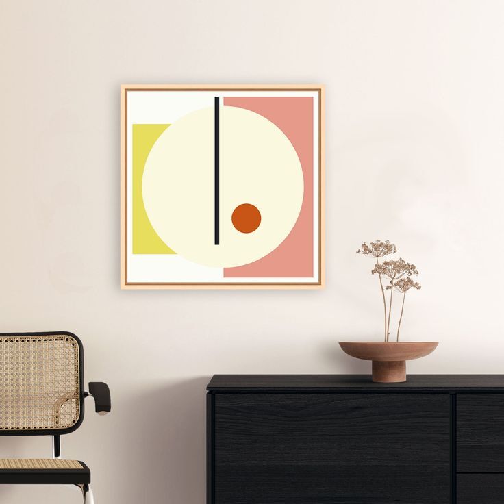 Quadratisches Leinwandbild mit abstrakten geometrischen Formen in Gelb, Weiß, Rosa und Schwarz, gerahmt und über einer schwarzen Kommode mit Trockenblumen in einer Vase und einem Stuhl platziert. Das Motiv besteht aus einem großen Kreis und einer vertikalen Linie, ergänzt durch eine kleine rote Kugel, im skandinavischen Stil gestaltet.