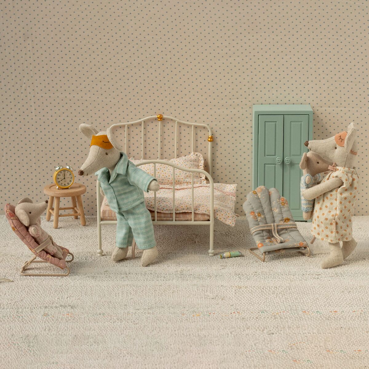 Zwei Stoffmäuse als Elternfigur und ein Stoffmausbaby in einer Zigarrenkiste, arrangiert in einem Miniaturschlafzimmer mit Bett, Schaukelstuhl, Spielzeuguhr, Miniaturschrank und pastellfarbenen Möbeln auf hellem, gepunktetem Hintergrund.