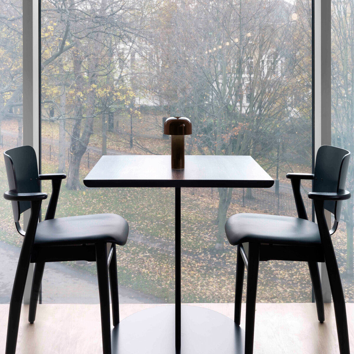 Eine moderne 'Bellhop' Tischleuchte auf einem Tisch neben zwei Stühlen vor einem großen Fenster, das eine Herbstlandschaft mit Bäumen zeigt.