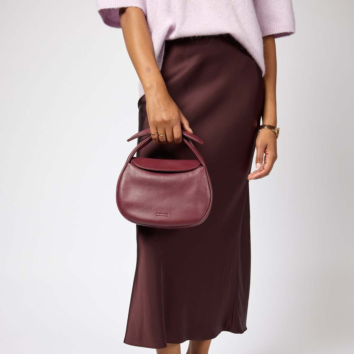 Eine Frau trägt eine elegante bordeauxrote Handtasche mit Henkel in Form eines Croissants, kombiniert mit einem passenden bordeauxroten Rock, fliederfarbenem Oberteil und dunkelroten Schuhen vor neutralem Hintergrund.
