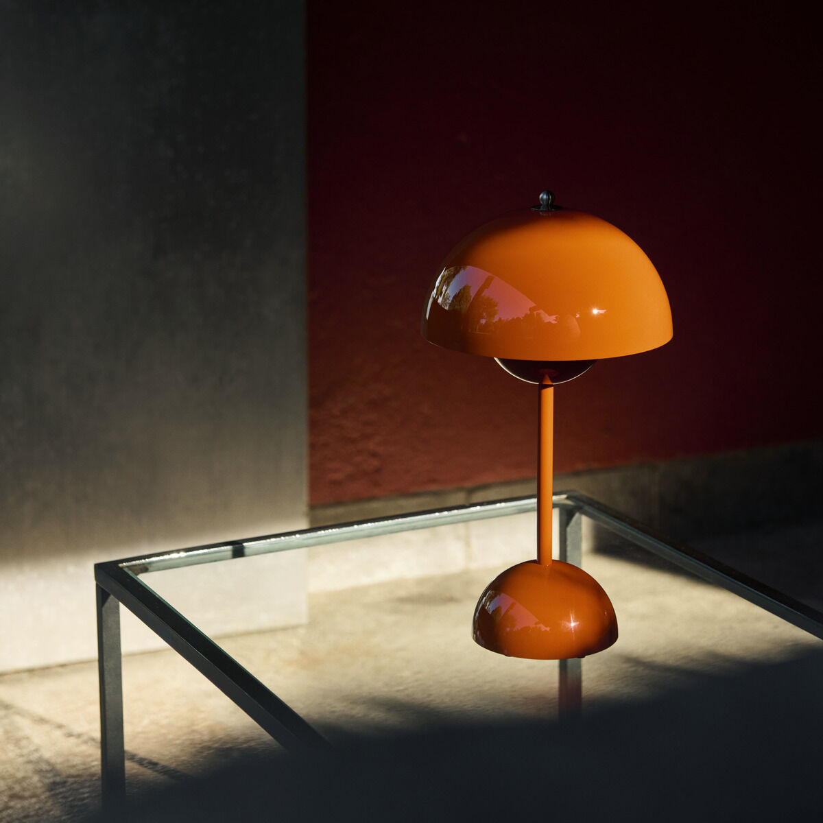 Die Flowerpot VP9 Tischleuchte Akku von &Tradition in der Variante Zesty Orange steht auf einem quadratischen Glastisch. Die orangefarbene Lampe im Retro-Design ist aus lackiertem Metall gefertigt. Im Hintergrund ist eine rote Wand mit Schattenwurf zu sehen, das Licht fällt gezielt auf die Leuchte.