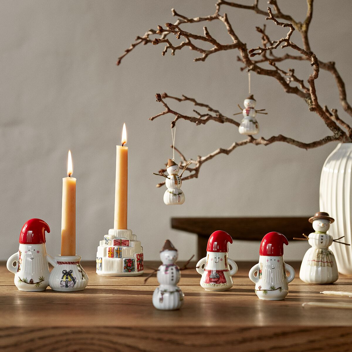 Mehrere dekorative Porzellanfiguren in Form von Weihnachtsmännern und Schneemännern aus der Hammershøi-Serie stehen auf einem Holztisch. Im Hintergrund brennen zwei Kerzen und von einem herbstlichen Zweig hängen weitere kleine Figuren.