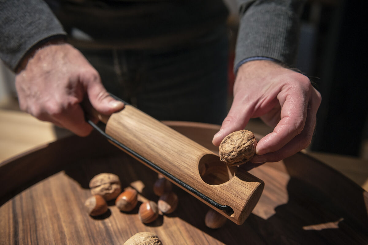Ein hölzerner Nussknacker mit dem Namen „Kracher“ wird verwendet, um eine Walnuss zu knacken. Mehrere ganze Walnüsse und Nüsse liegen in einer Holzschale. Eine Person hält die Nuss und den Nussknacker in den Händen.