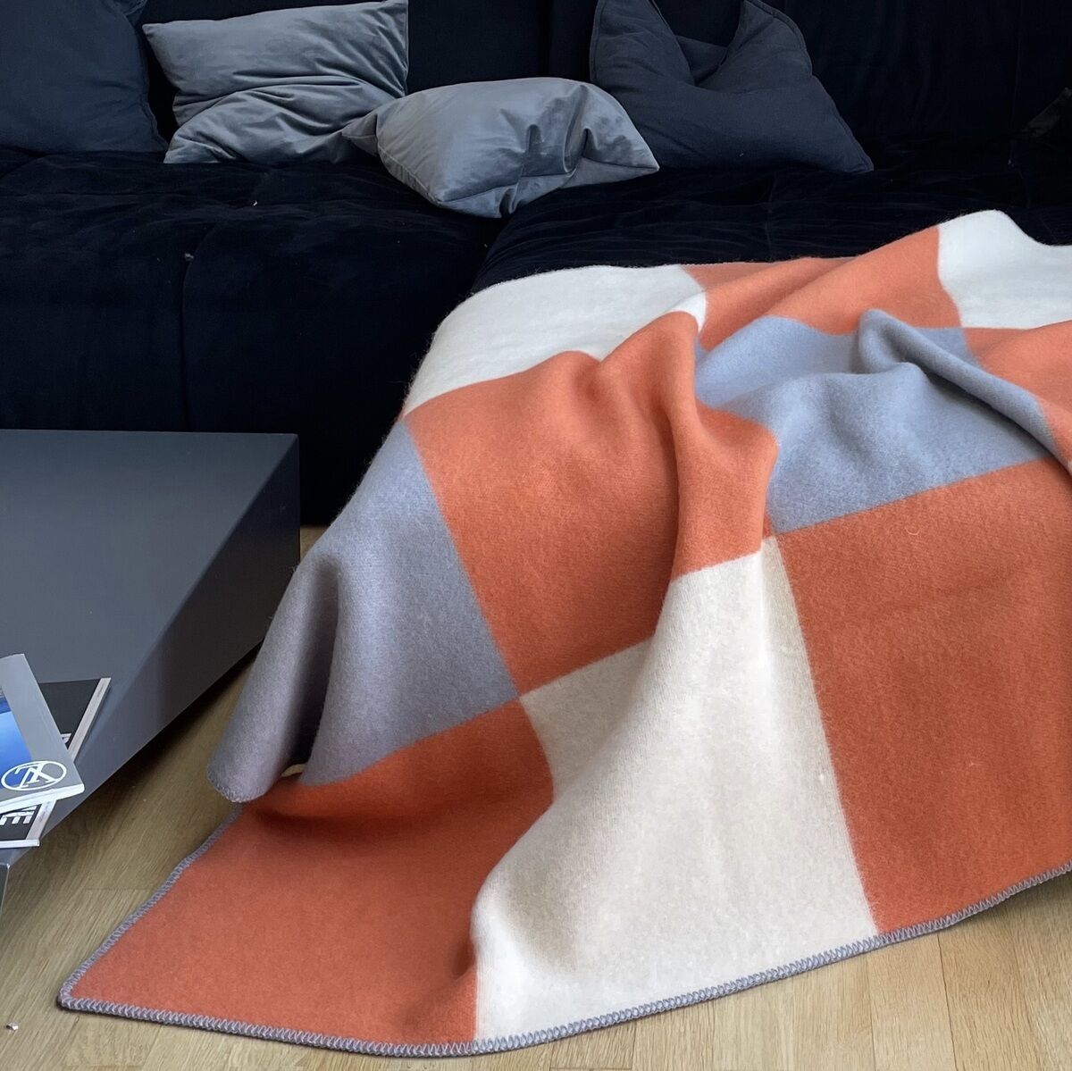 Ein modernes Wohnzimmer mit einem schwarzen Sofa, dekoriert mit einer Tricolor-Decke im Karomuster. Die Decke ist in den Farben Orange, Grau und Beige gehalten. Im Hintergrund hängt ein großes Bild von Paris und eine schwarze Stehlampe ragt über das Sofa.