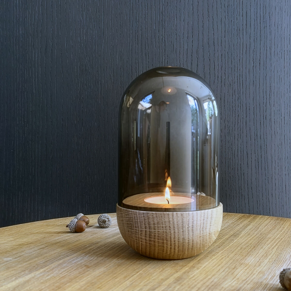 Windlicht »GLOW« von finbeaux auf hellem Holztisch. Es besteht aus einer runden Holzbasis und einem rauchfarbenen, gläsernen Aufsatz, unter dem eine brennende Kerze steht. Umgeben von Eicheln, vor dunkelgrauer Wand, stilvoll und modern arrangiert.