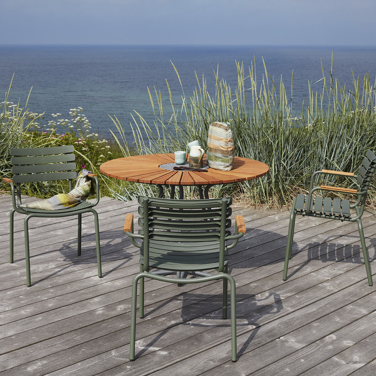 Auf einer Holzterrasse stehen drei grüne Gartenstühle um einen runden Holztisch. Der Hintergrund zeigt das Meer und das Küstenland mit hohem Gras und Blumen.