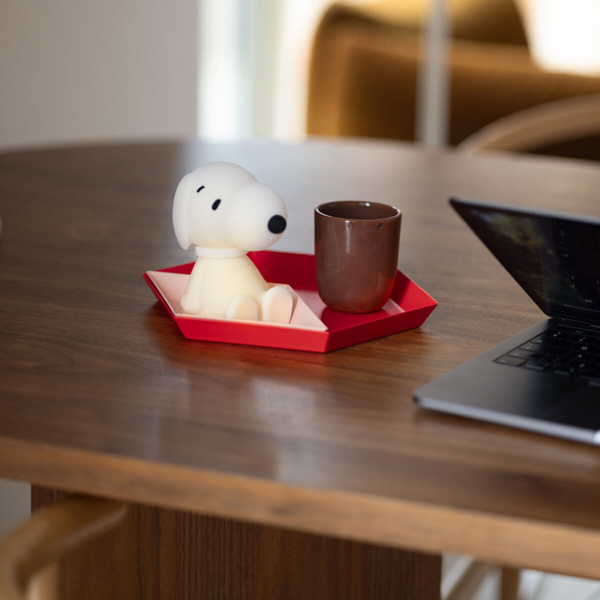 Ein Nachtlicht in Form von Snoopy steht auf einem sechseckigen, roten Tablett neben einer braunen Tasse auf einem Holztisch mit Laptop; im Hintergrund unscharfe Wohnzimmereinrichtung und Tageslicht.