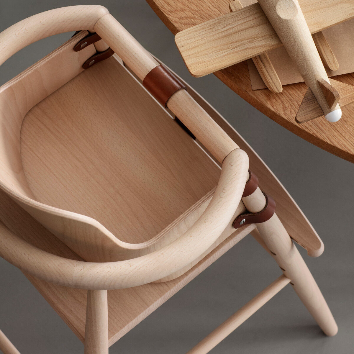 Ein hochwertiger Baby-Einsatz aus Holz für einen Kinderhochstuhl ist auf einem Tisch abgebildet. Neben dem Einsatz liegt ein kleines Holzflugzeug. Die Materialien wirken nachhaltig und ergonomisch designt.
