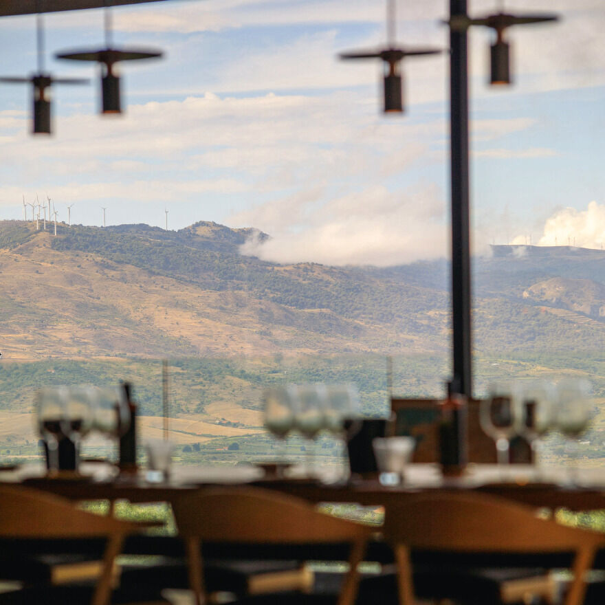 Im Vordergrund ein elegant gedeckter Tisch mit Weingläsern und Flaschen, dahinter große Panoramafenster mit Blick auf eine hügelige, grüne und braune sizilianische Landschaft unter blauem Himmel mit vereinzelten Wolken und Windrädern auf einem Bergrücken.