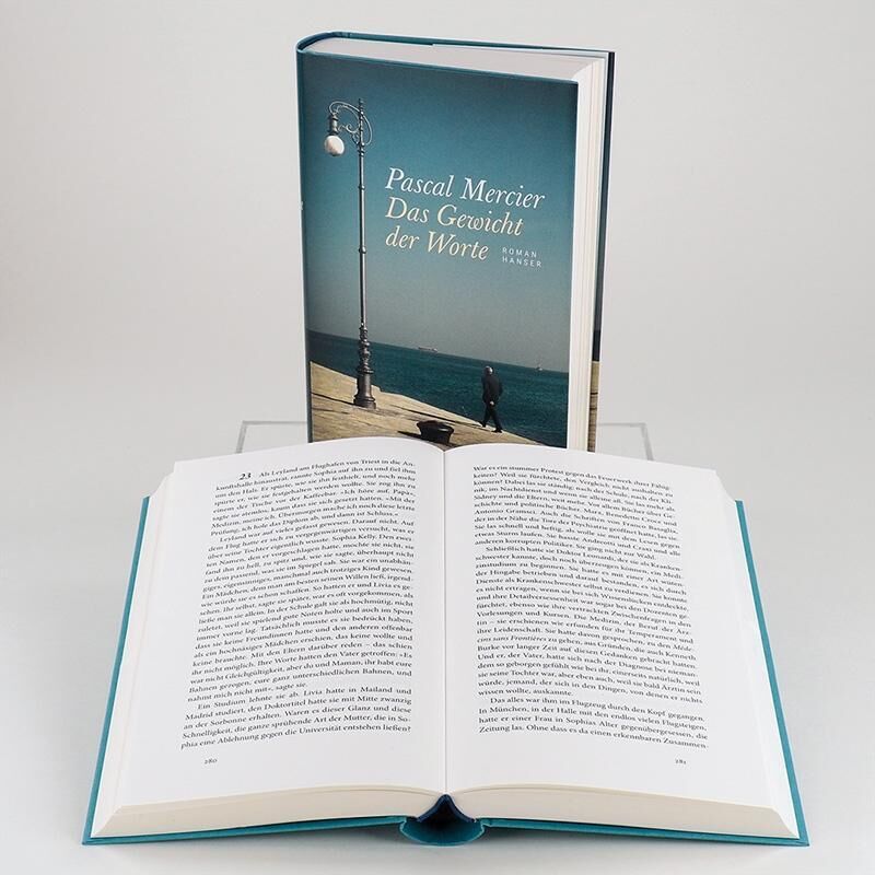 Ein aufgeschlagenes Buch steht vor einem aufrecht stehenden Buch mit dem Titel 'Pascal Mercier: Das Gewicht der Worte'. Das Cover zeigt einen Spaziergang am Meer unter blauem Himmel.