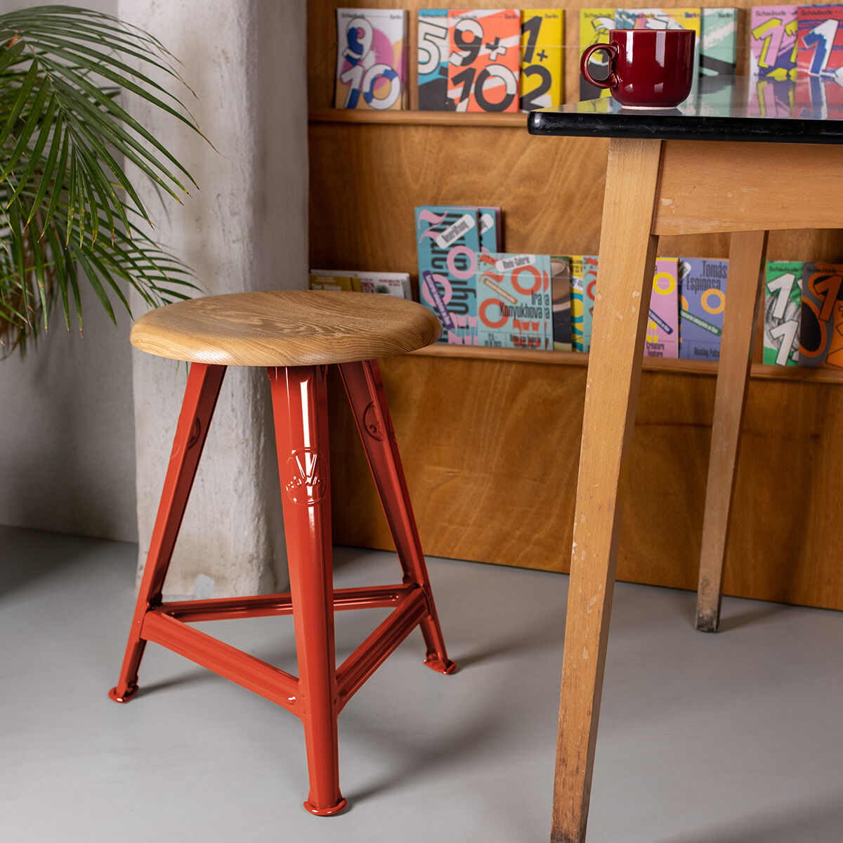 Ein roter Metallhocker mit Holzsitzfläche steht neben einem Tisch im Industriestil. Im Hintergrund befindet sich eine Regalwand mit bunten, nummerierten Deko-Elementen. Pflanzen ergänzen das moderne, künstlerische Ambiente des Raumes.