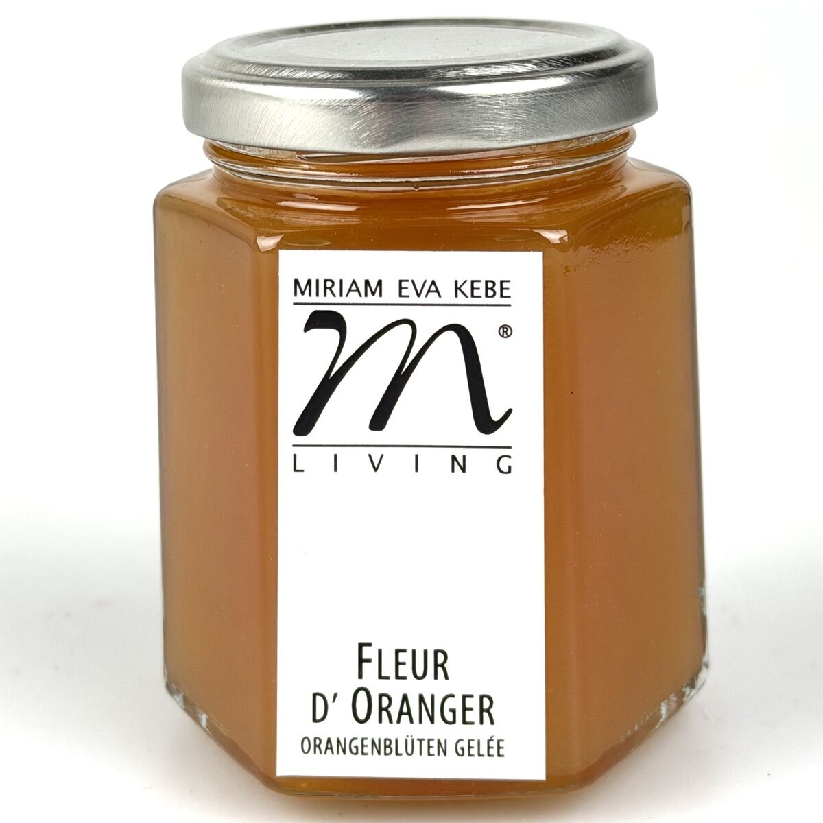 Sechseckiges Glas mit silbernem Deckel, gefüllt mit orangefarbenem Orangenblüten-Gelee. Auf dem weißen Etikett steht "Miriam Eva Kebe Living" und "Fleur d'Oranger Orangenblüten Gelee" in schwarzer Schrift.