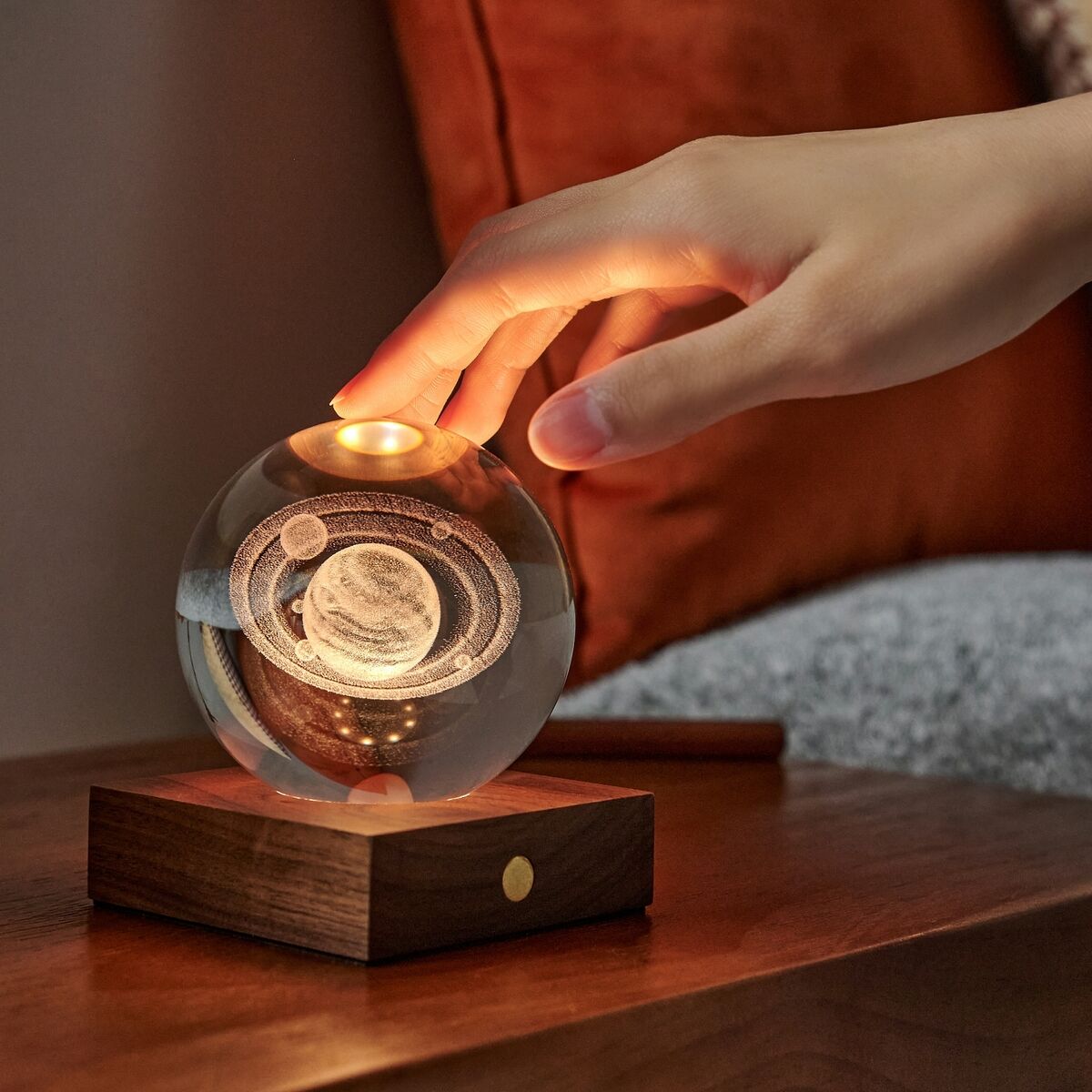 Eine moderne Lampe namens »Amber Light« steht auf einem Holztisch im Wohnzimmer. Die Lampe besteht aus einer leuchtenden Glaskugel, die auf einer schlichten Holzbasis ruht, während eine Hand sie sanft berührt. Im Hintergrund liegen orangefarbene und graue Sofakissen. Das warme Licht erzeugt eine gemütliche Atmosphäre.