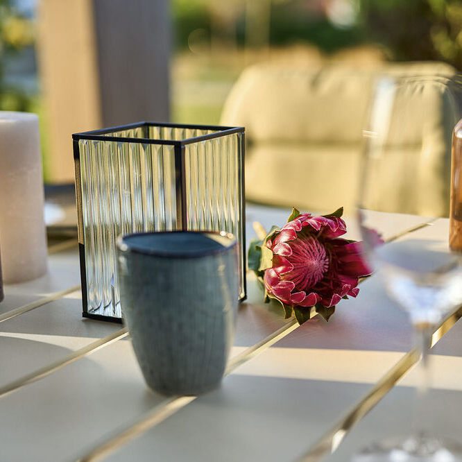 Ein gedeckter Tisch im Garten mit zwei Weingläsern, einer Kerze, einer Vase mit einer Blume, einer Tasse und zwei Flaschen Olivenöl auf einer beigen Tischplatte.