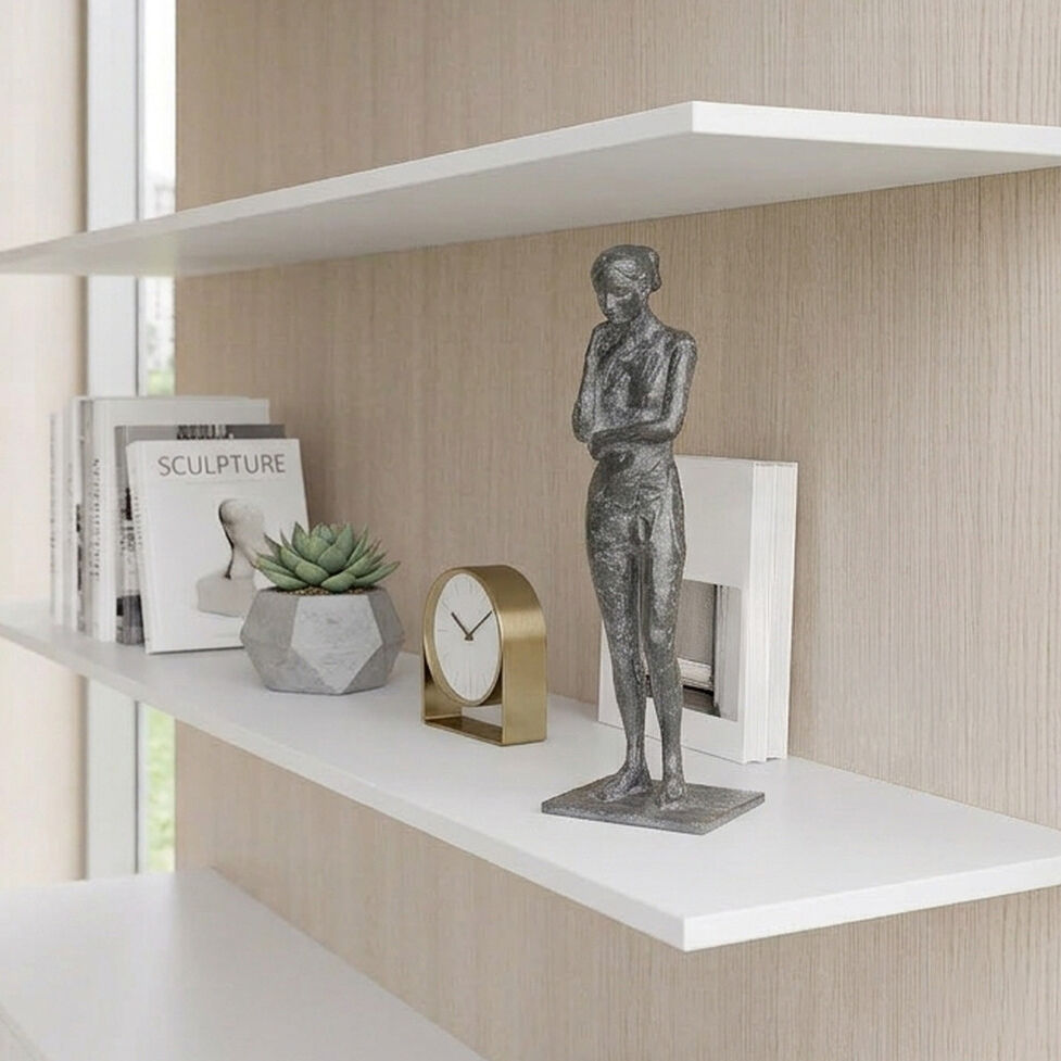 Auf einem weißen Regal steht die Bronze-Skulptur »Besinnung« von Valerie Otte. Sie zeigt eine schlanke, stehende Frauenfigur mit verschränkten Armen. Daneben stehen eine goldene Tischuhr, ein geometrischer Blumentopf mit Sukkulente und Bücher, moderner Wohnstil, helles Ambiente.