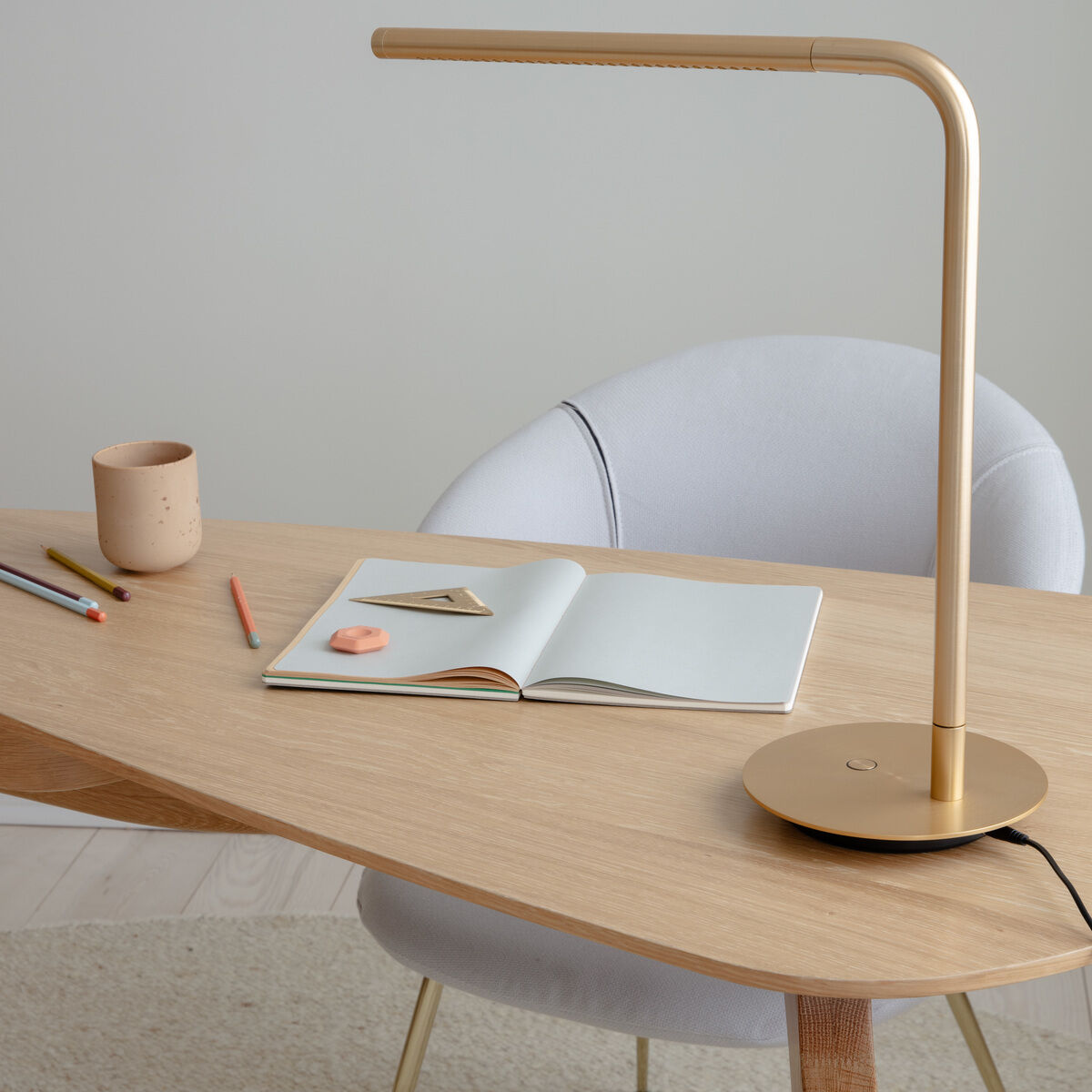 Moderner, heller Schreibtisch mit abgerundeten Kanten aus Holz, darauf eine goldfarbene, minimalistische Tischlampe, ein geöffnetes Notizbuch, einige bunte Stifte und eine beige Tasse. Der Schreibtisch steht auf hellem Holzboden und wird von einem gepolsterten hellgrauen Stuhl begleitet. Die Szene vermittelt Ordnung, Kreativität und eignet sich ideal für Arbeits- oder Lernumgebungen.