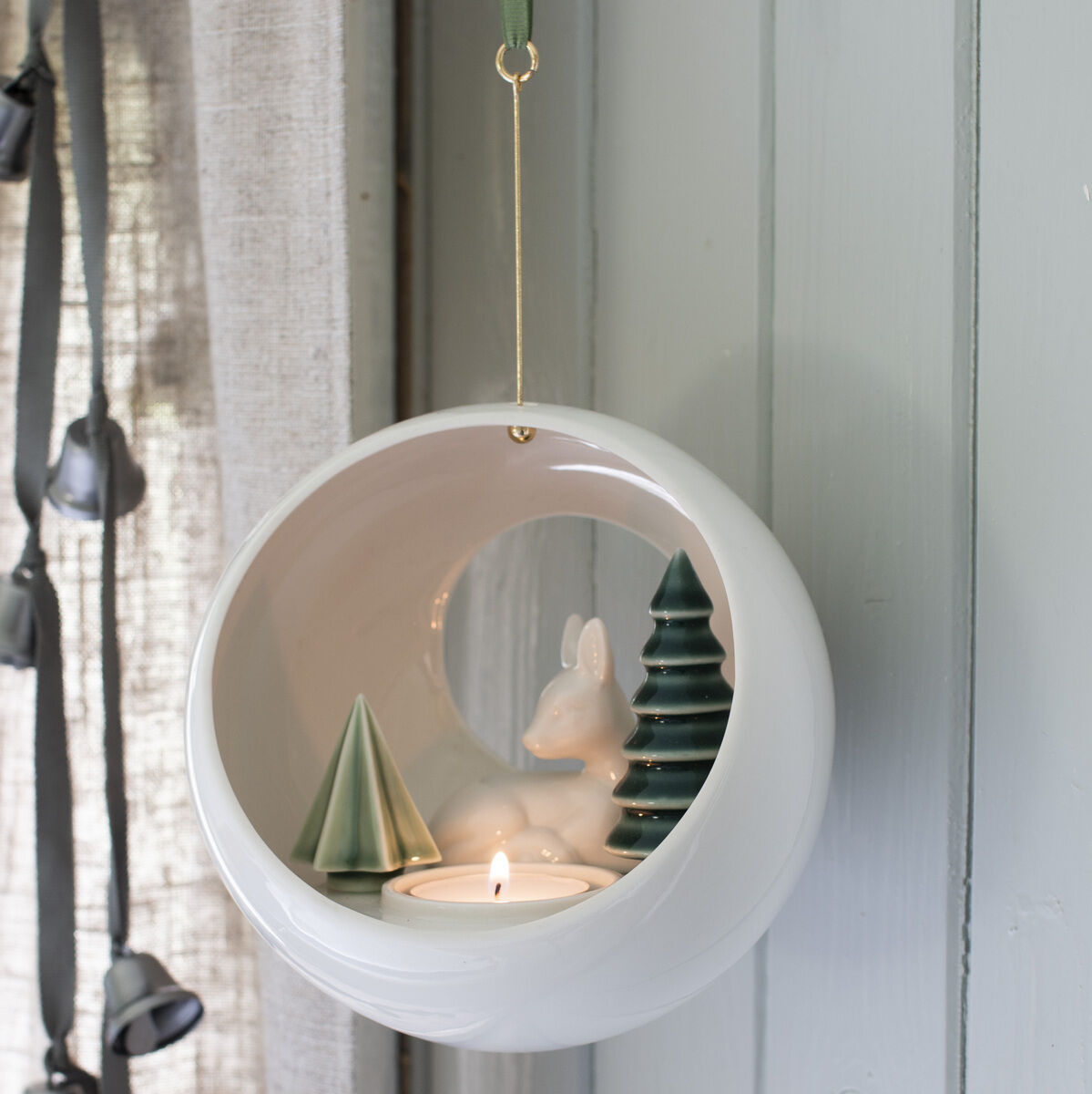 Ein weißer, runder Baumschmuck mit hängendem Kerzenhalter. Im Inneren des Kreises befinden sich ein Porzellan-Rehkitz und zwei grüne Tannen. Der Schmuck hängt an einem grünen Band und einem goldenen Faden.