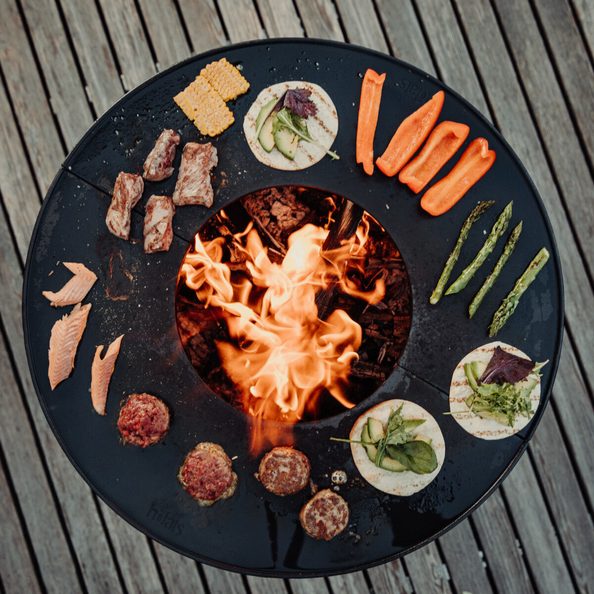 Ein rundes Grillset auf einem Holzboden mit verschiedenen gegrillten Lebensmitteln wie Gemüse, Fleisch und Fisch um eine zentrale Feuerstelle herum platziert.