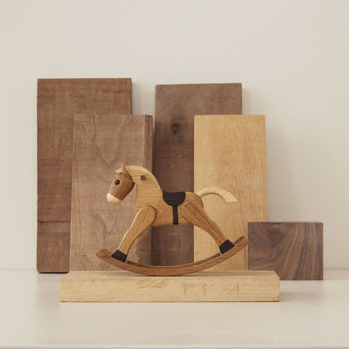 Eine handgefertigte Holzfigur in Form eines Schaukelpferds steht vor mehreren aufrecht gestellten Holzplatten. Das detailreiche Schaukelpferd besteht aus verschiedenen Naturholztönen und dient als dekoratives Element oder Spielzeug, ideal für Kinder oder als traditionelle Dekoration.