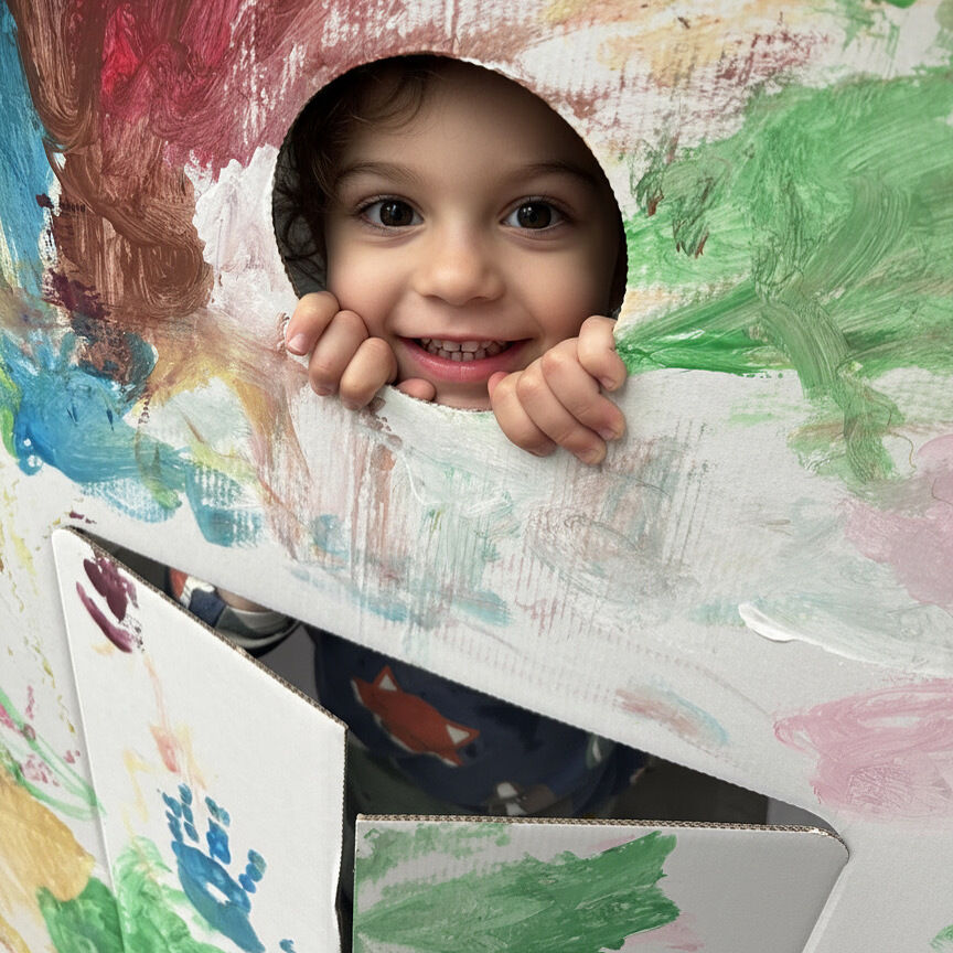 Ein lachendes Kind blickt durch das runde Fenster eines großen, bemalten Papphauses. Das Haus aus weißer Pappe ist bunt mit Fingerfarben bemalt. Das Kind hält sich an der Kante fest und wirkt fröhlich und kreativ beschäftigt.