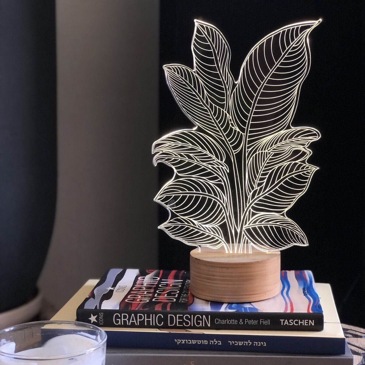 3D-Lampe im Heliconia-Design mit Holzsockel, beleuchtet auf einem Stapel Bücher neben einer weißen Kerze, im Hintergrund ein schwarzer Vorhang.