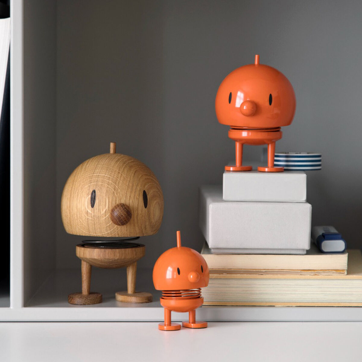 Ein Regal mit mehreren Hoptimist Bumble Figuren aus Eiche und orangefarbenem Kunststoff. Die Figuren stehen zwischen Bücherstapeln und dekorativen Elementen. Eine größere Holzfigur und zwei kleinere orangefarbene Figuren sind sichtbar. Sie strahlen Freude und Optimismus aus.