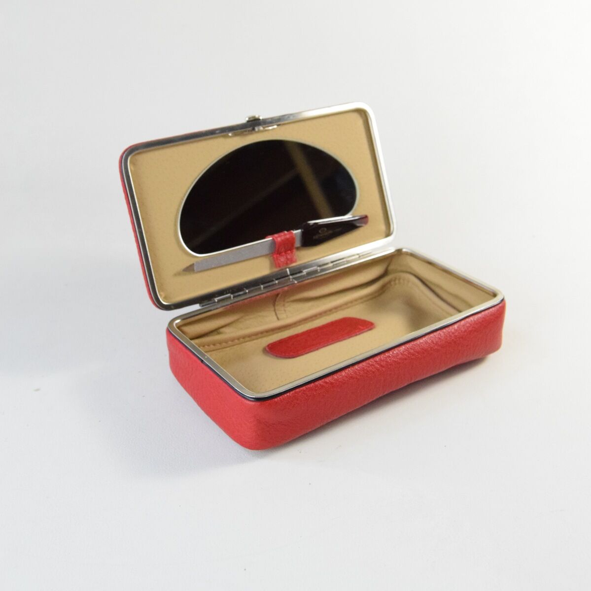 Eine geöffnete Utensilienbox aus rotem Leder mit Messingbeschlägen. Die Box hat ein beige-farbenes Inneres und enthält einen integrierten Spiegel sowie eine Halterung für einen Stift.