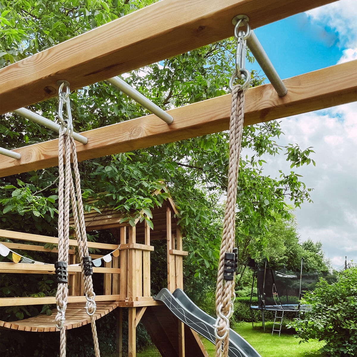 Ein Holz-Spielplatz mit einer Skateboard-Schaukel, Seilen und einer Rutsche im Garten bei sonnigem Wetter.