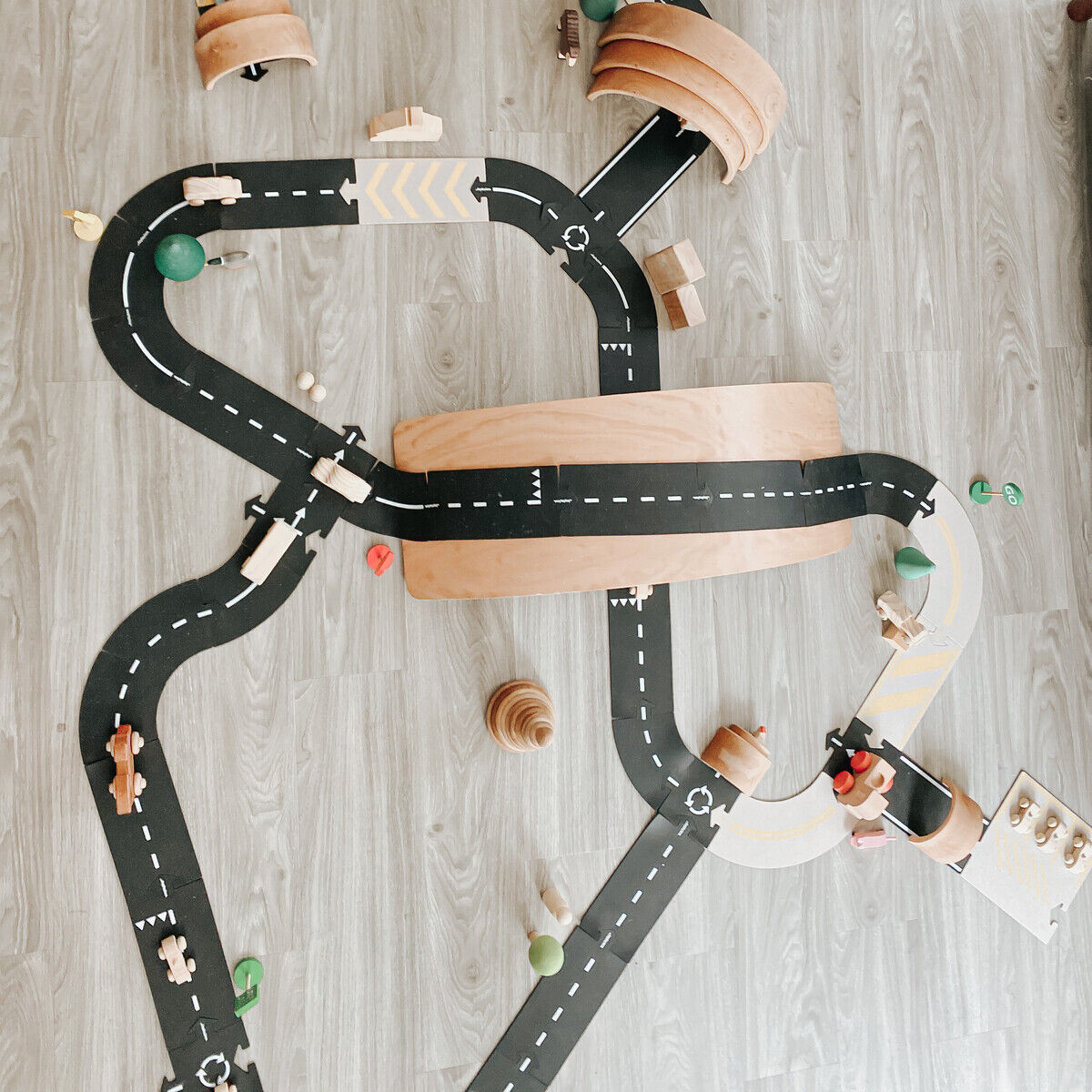 Kind spielt auf hellgrauem Boden mit einem großen Straßenbau-Set aus Holz und schwarzen Fahrbahn-Teilen. Verschiedene Kurven und Brücken sind aufgebaut; einige Holzfahrzeuge und kleine Spielfiguren ergänzen die Szene. Das Set fördert kreatives Bauen und Rollenspiele.