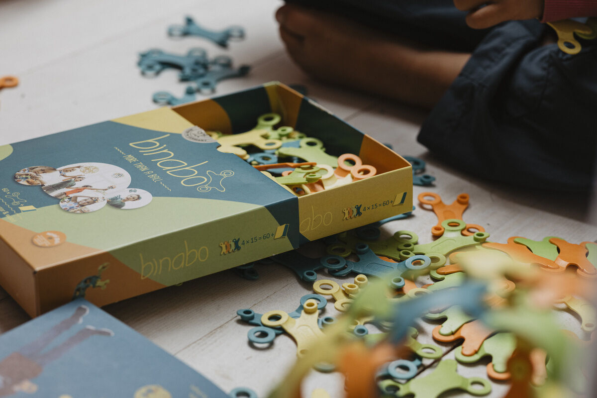 Spiel »Binabo« 60 Teile - Bunte, recycelbare Kunststoffteile in einer offenen Schachtel, umgeben von Kinderhänden, die darauf hinweisen, dass sie bald zum Spielen verwendet werden.